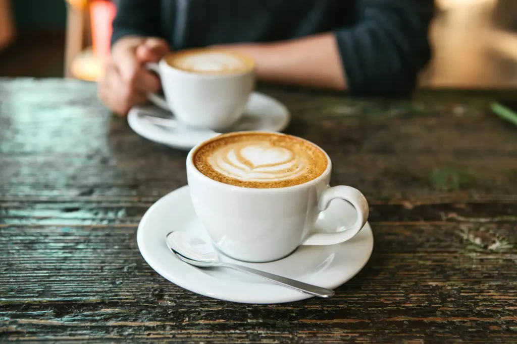 Two cups of coffee on saucers sit on a rustic wooden table, with one cup featuring latte art and a person holding the other.