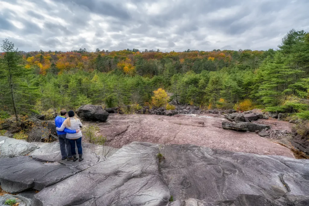A couple stands on rocky terrain overlooking a river and a forest with colorful autumn foliage under a cloudy sky.