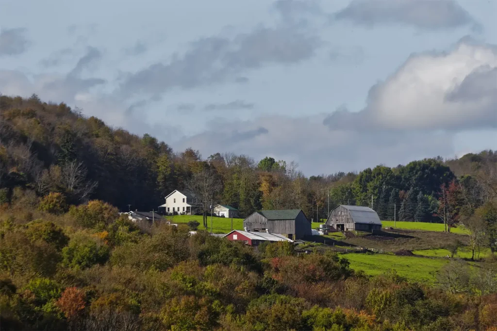 Rolling hills with a cluster of farm buildings and a white house surrounded by lush greenery and autumn foliage under a partly cloudy sky.