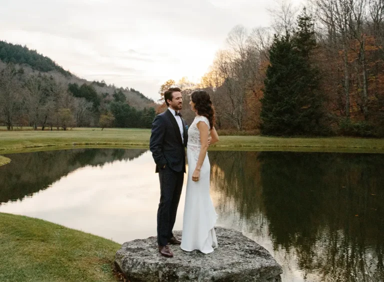 A couple stands on a rock by a serene pond during sunset, surrounded by trees and mountains.