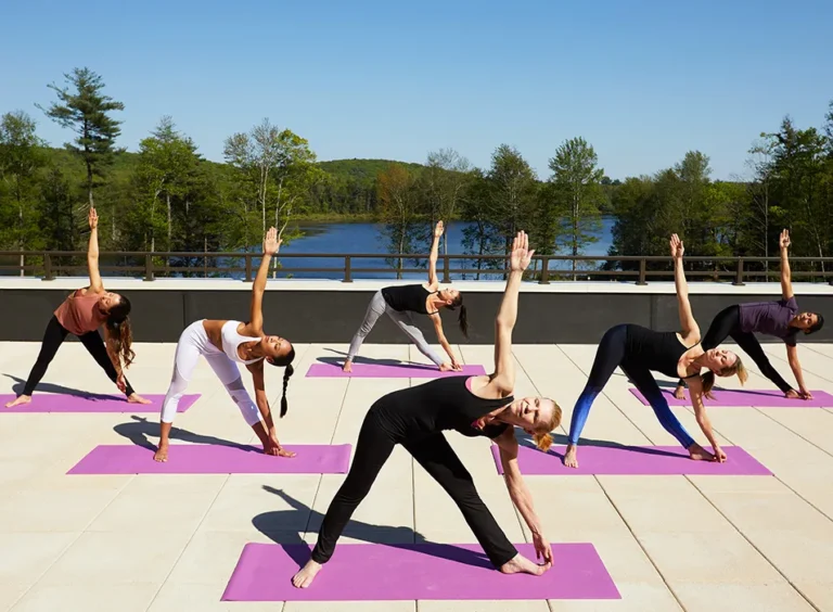 A group of women practicing yoga outdoors on a sunny day, with a scenic lake and trees in the background.