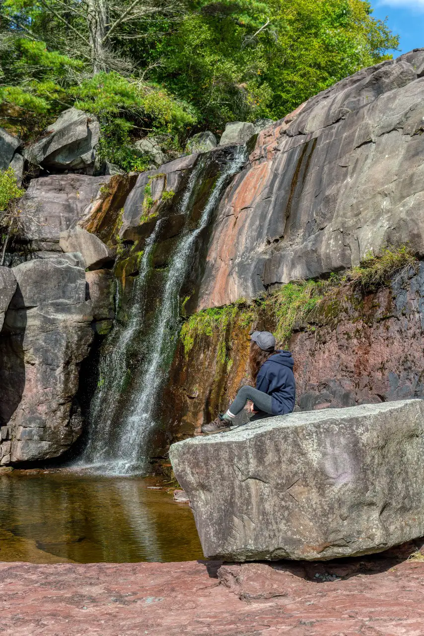 A tranquil scene of a person sitting on a rock near a small waterfall surrounded by lush greenery.