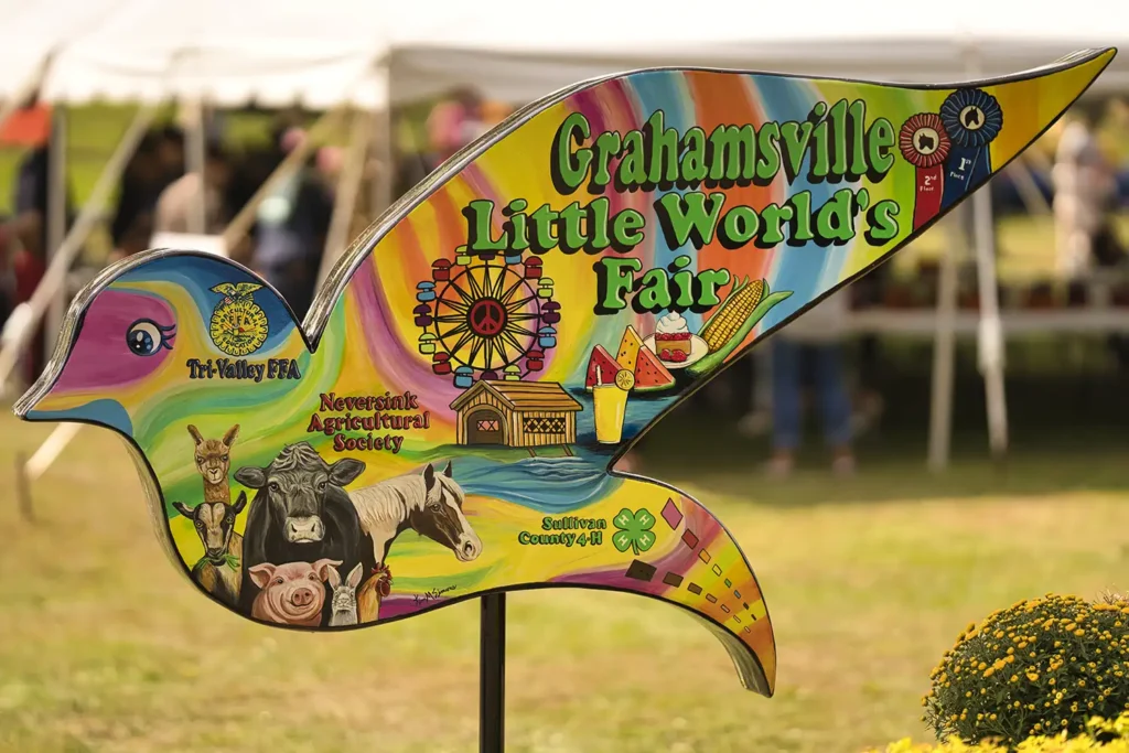A colorful sign shaped like a bird, advertising the Grahamsville Little World's Fair, featuring various fair-themed illustrations.
