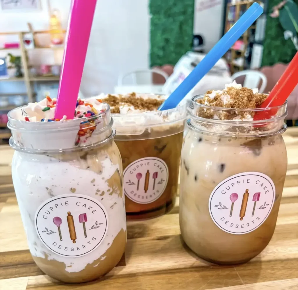 Three dessert jars filled with colorful drinks topped with whipped cream and crumbs, featuring vibrant straws and branded labels.