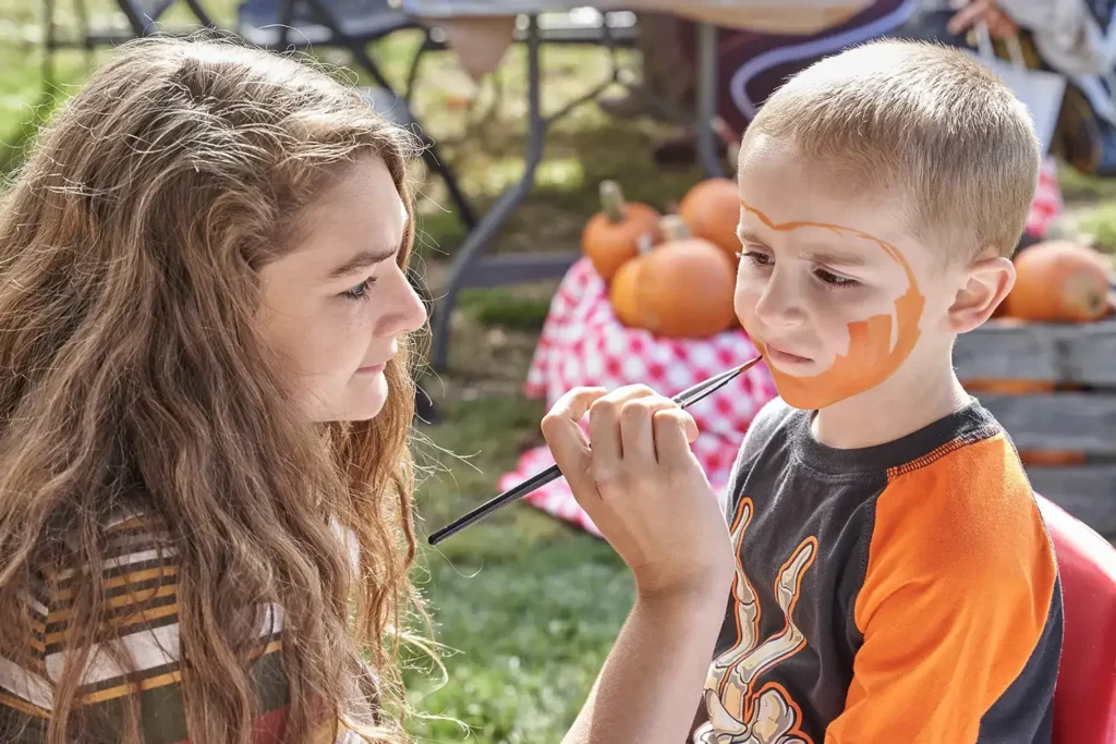 A girl paints a boy's face at an outdoor event, surrounded by pumpkins and a festive atmosphere.