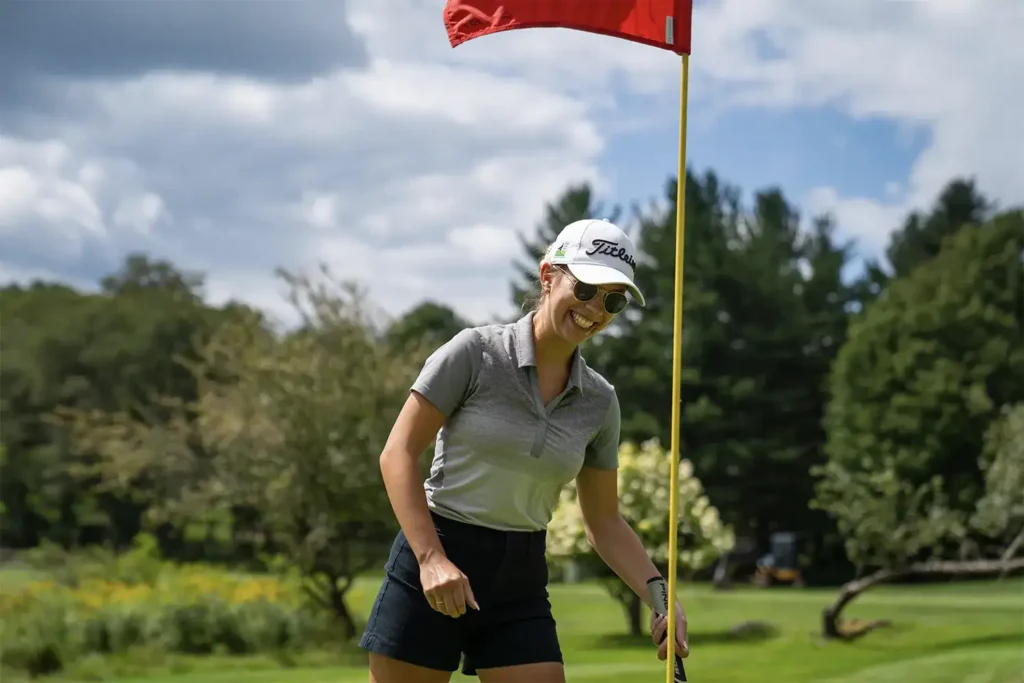 A woman smiles near a golf flag on a green course, dressed in casual golf attire, with trees and clouds in the background.