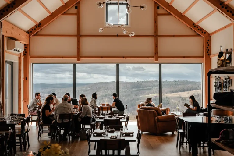 A spacious restaurant interior with wooden beams, large windows offering scenic views, and patrons enjoying meals and drinks.