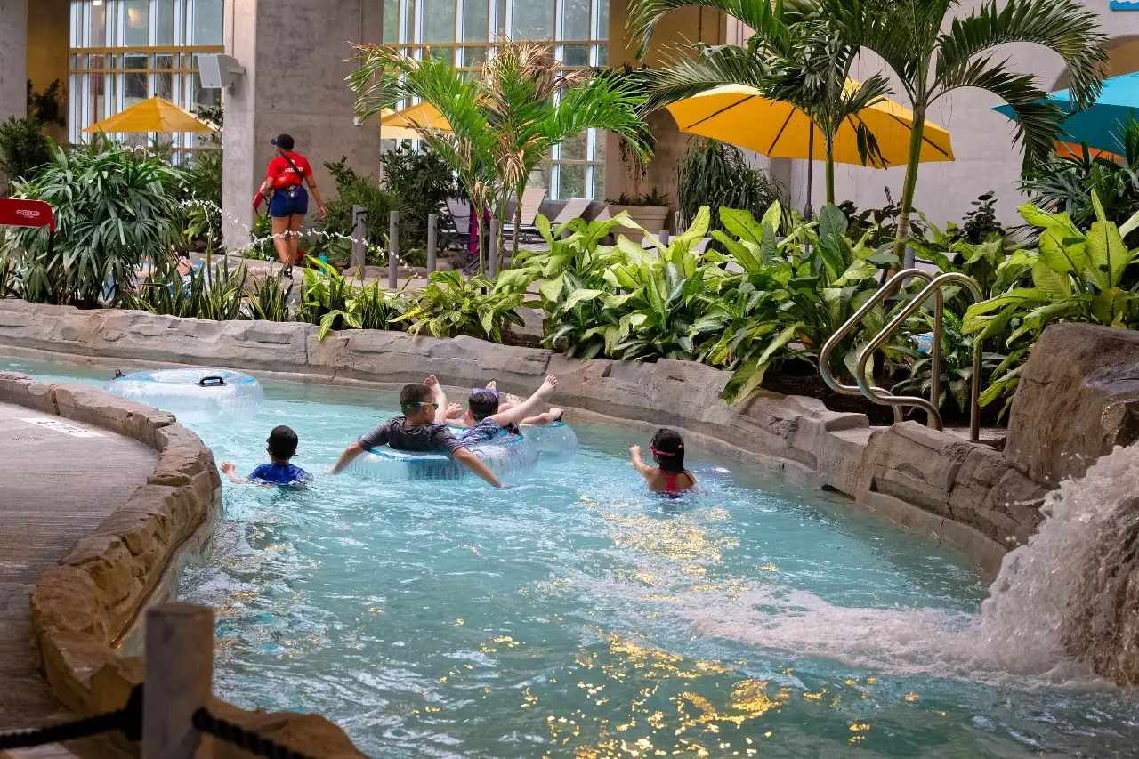 A tropical indoor pool area with guests enjoying the water, surrounded by lush greenery and yellow umbrellas.