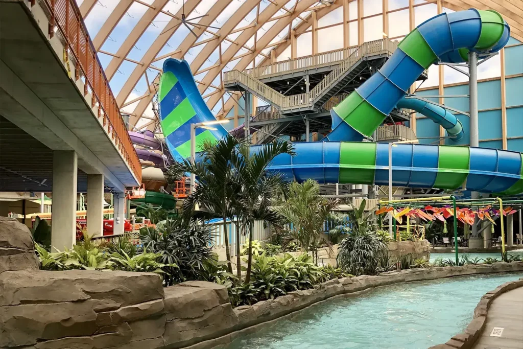 An indoor water park featuring a large, colorful waterslide, tropical plants, and a lazy river surrounded by decorative rocks.