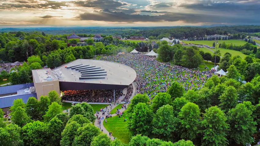 A large crowd gathers around an outdoor concert venue surrounded by trees, with a scenic sky in the background.