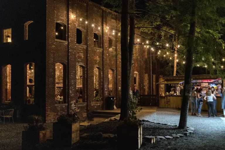 An outdoor gathering spot at night, featuring string lights, a rustic building, and groups of people enjoying themselves.