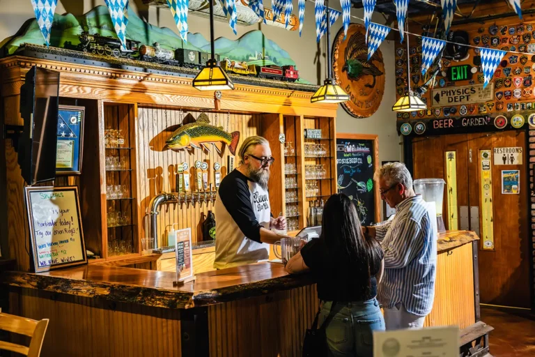 A bar scene with a bartender serving drinks to two customers, surrounded by decorations and memorabilia in a lively atmosphere.