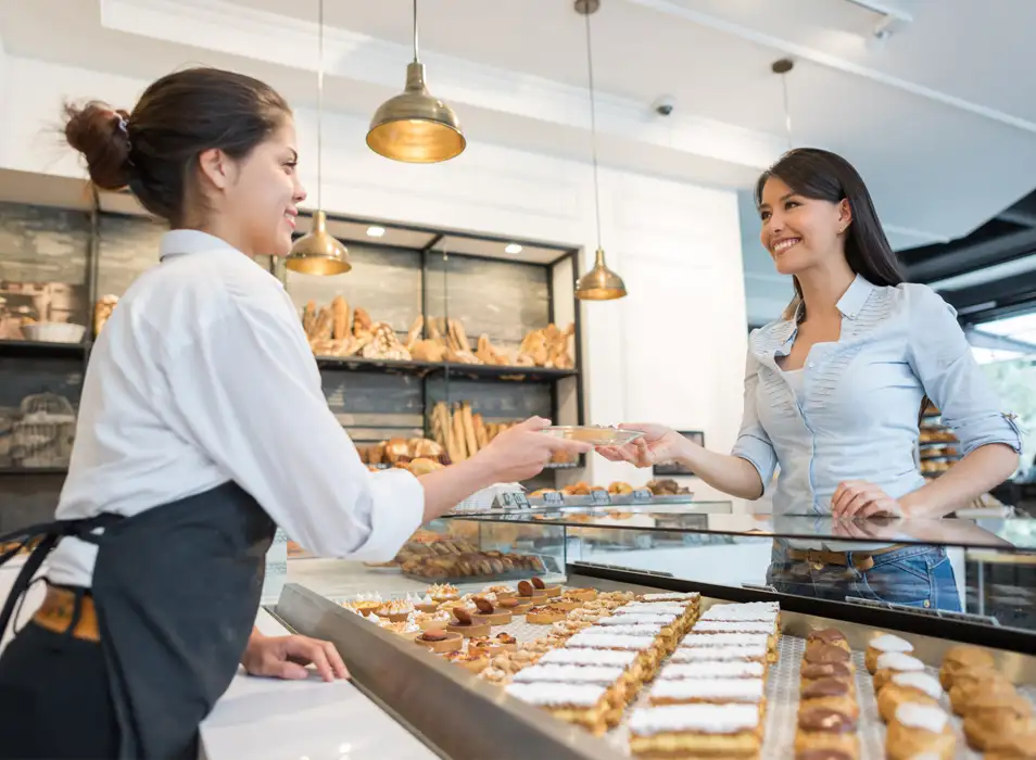 A bakery scene with two women; one in an apron serves a customer, while a selection of pastries is displayed prominently.