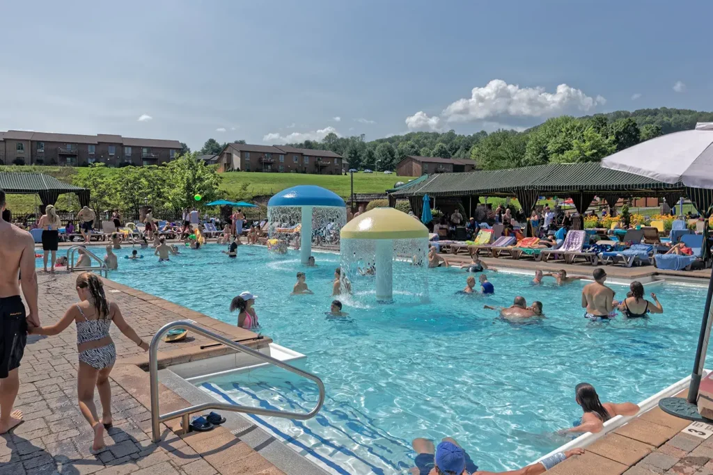 A lively pool area features children and adults enjoying the water, with colorful mushroom-shaped fountains and lounge chairs under a sunny sky.