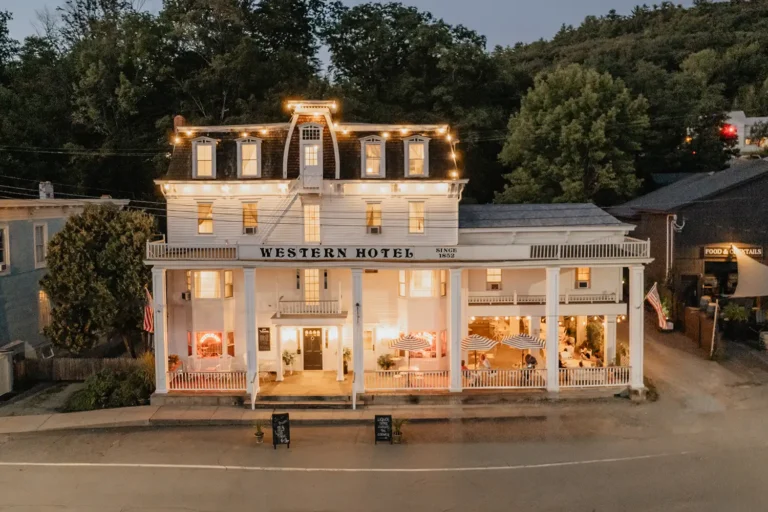 A charming hotel with festive lighting, featuring a classic facade and outdoor seating, surrounded by greenery and a calm street atmosphere.