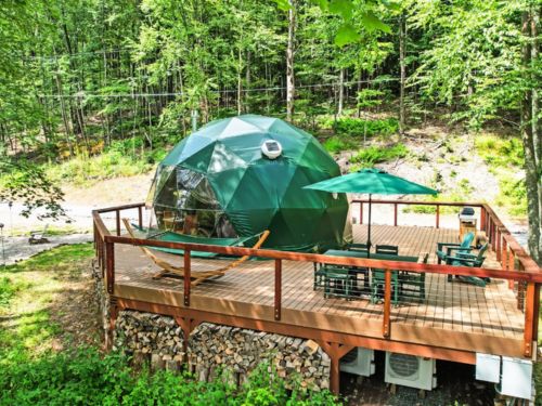 A green geodesic dome cabin on a wooden deck, surrounded by lush forest, featuring patio furniture and a serene outdoor setting.