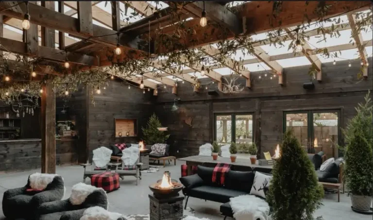 A cozy indoor space with wooden decor, greenery, and string lights, featuring comfortable seating and a warm fire pit.
