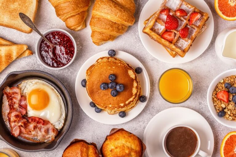 A breakfast spread including pancakes, bacon, croissants, waffles, fresh fruits, and beverages like orange juice and coffee.