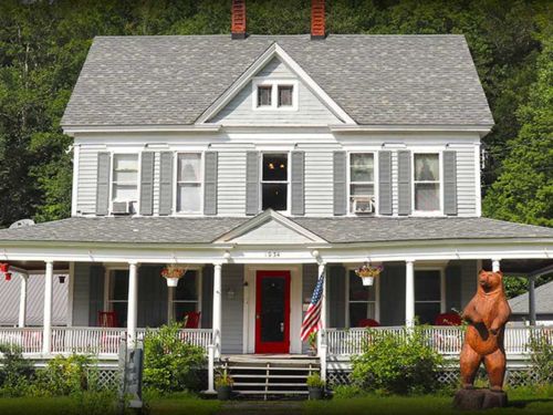 A charming gray Victorian house with white trim, a red door, and a wooden bear statue in the front yard, surrounded by greenery.