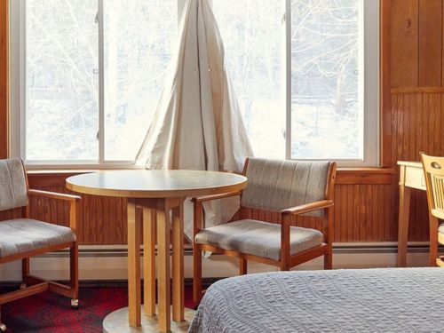 A cozy room with a wooden table and chairs, featuring a large window with a curtain and a bed in the foreground.