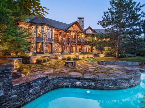 A stunning stone mansion with multiple stories, surrounded by lush greenery and a serene pool, illuminated by soft evening lights.