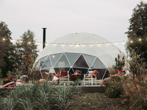 A geodesic dome structure surrounded by lush greenery, decorated with lights and outdoor seating, creating a cozy, inviting atmosphere.