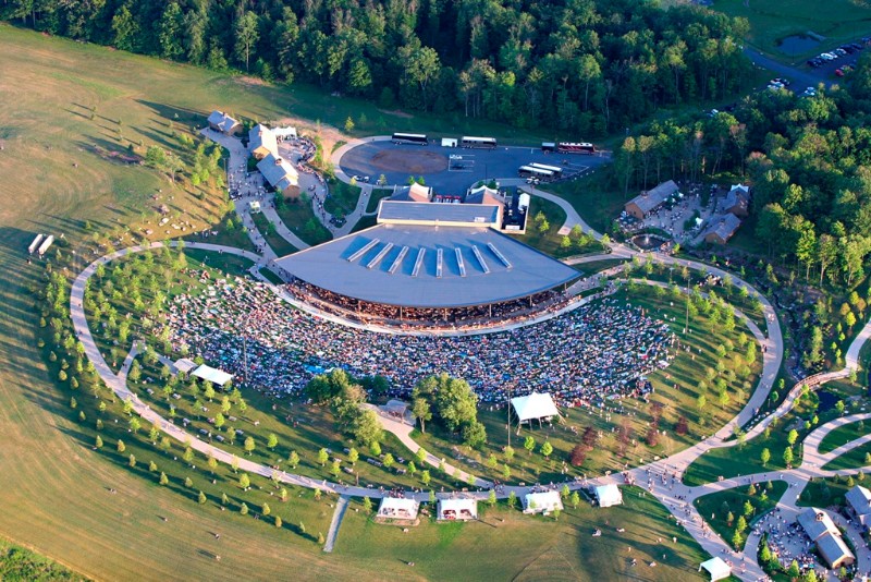 An aerial view of a large outdoor concert venue filled with people, surrounded by lush greenery and pathways.