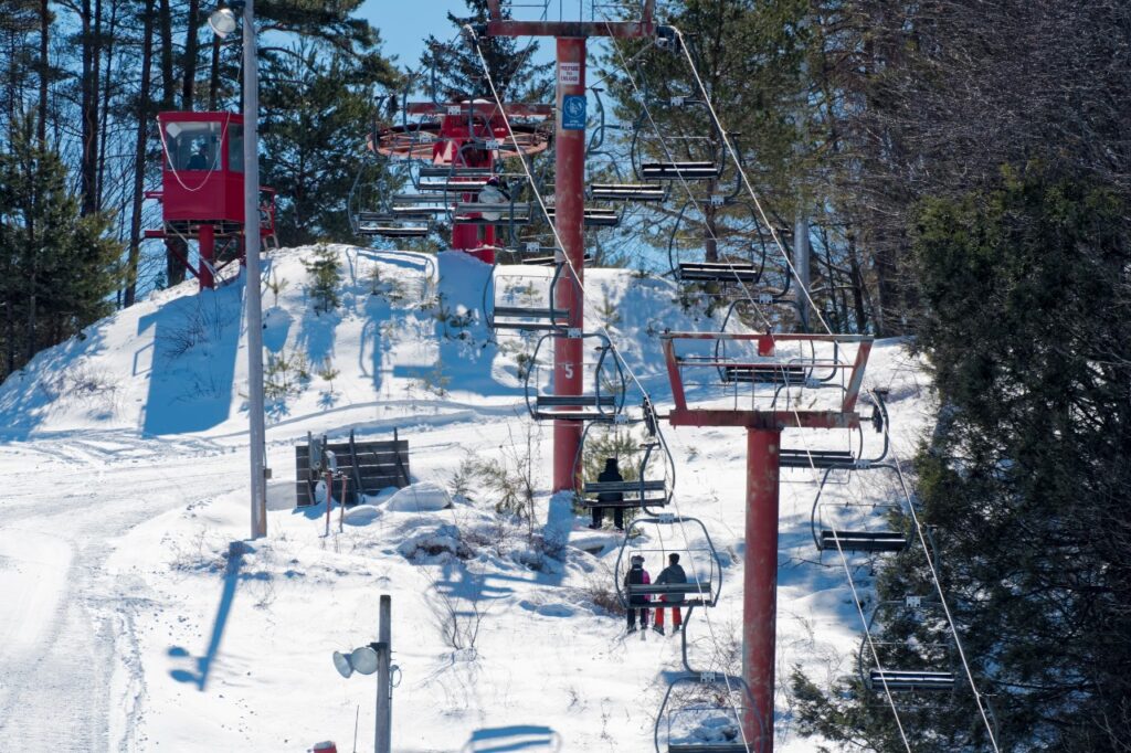 Skiers and snowboarders ride a chairlift up Holiday Mountain, surrounded by forest and snow trails underneath them.