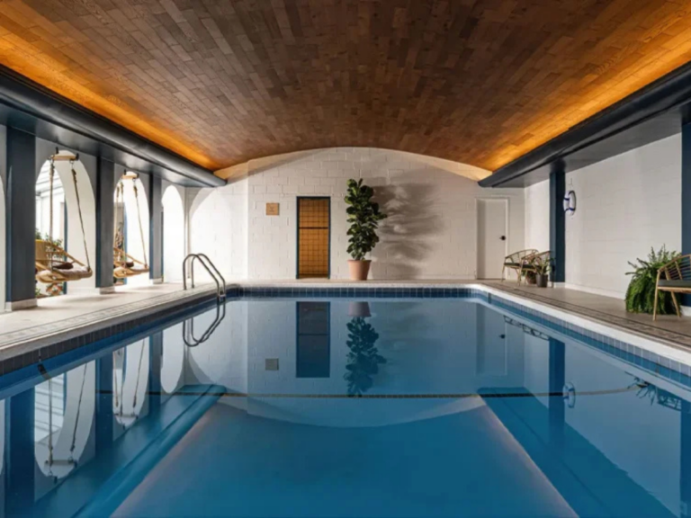 A serene indoor swimming pool with a modern design, featuring wooden ceilings, elegant lighting, and large arched windows.