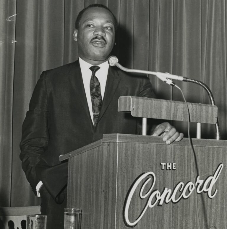A man stands at a podium labeled 'The Concord', delivering a speech, dressed in formal attire with a confident expression.
