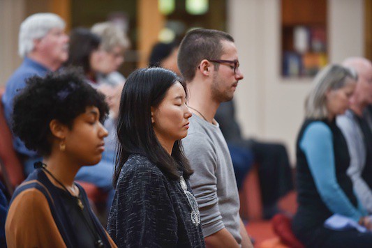 A group of individuals sits in a meditative posture, focusing inwardly in a serene environment, with soft lighting and a calm atmosphere.