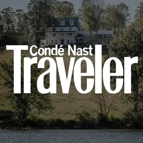 A serene landscape featuring a house surrounded by trees, with a lake in the foreground, highlighting the text "Condé Nast Traveler."