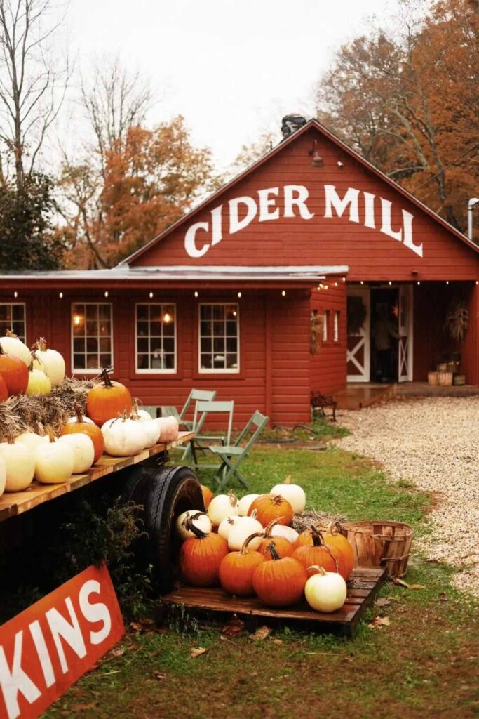 A picturesque cider mill with a bright red exterior, surrounded by various pumpkins, set against a backdrop of autumn foliage.