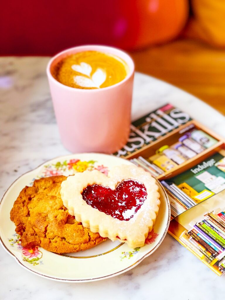 A cozy scene featuring a pink cup of coffee, a heart-shaped cookie with jam, a chocolate chip cookie, and magazines on a marble table at 2 Queens Coffee.