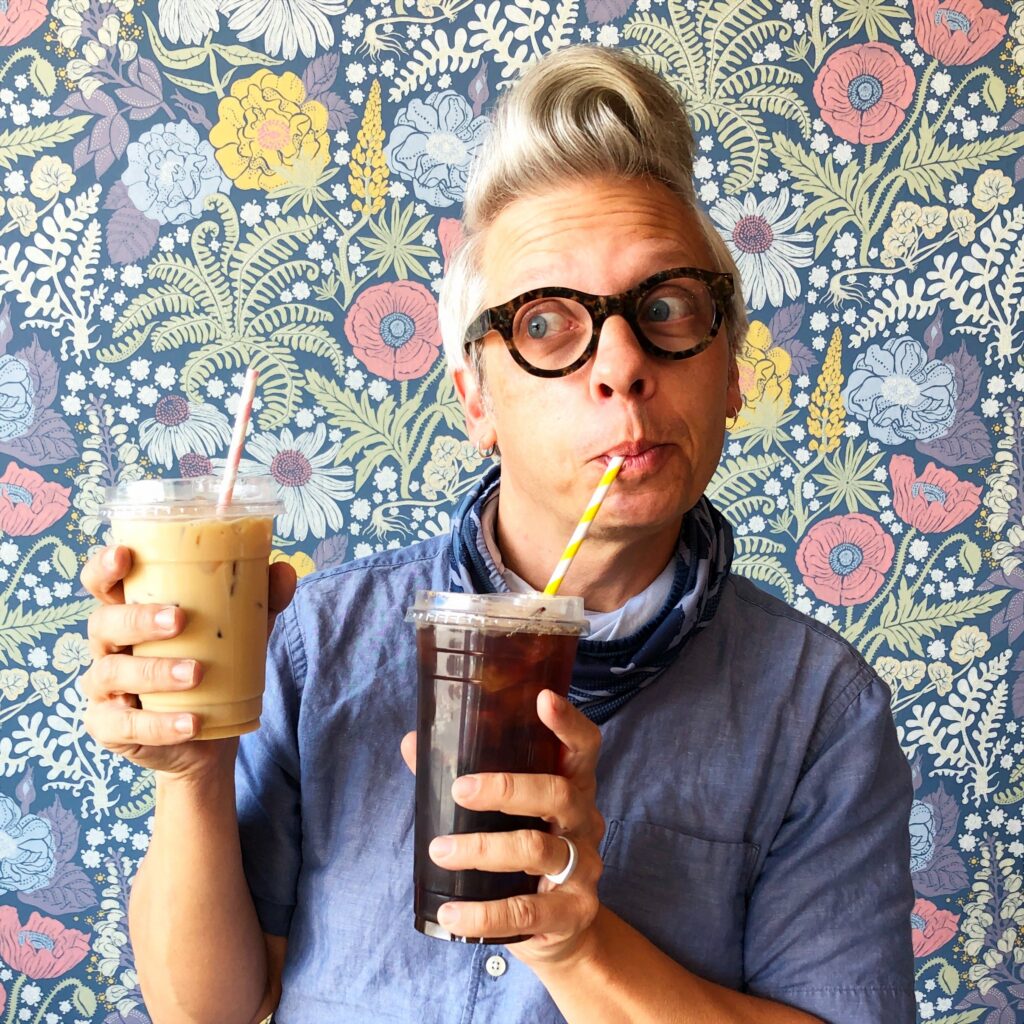 Charles Wilkin, co-owner of 2 Queens Coffee, holding two drinks, one iced coffee and one cold brew, against a colorful floral background.