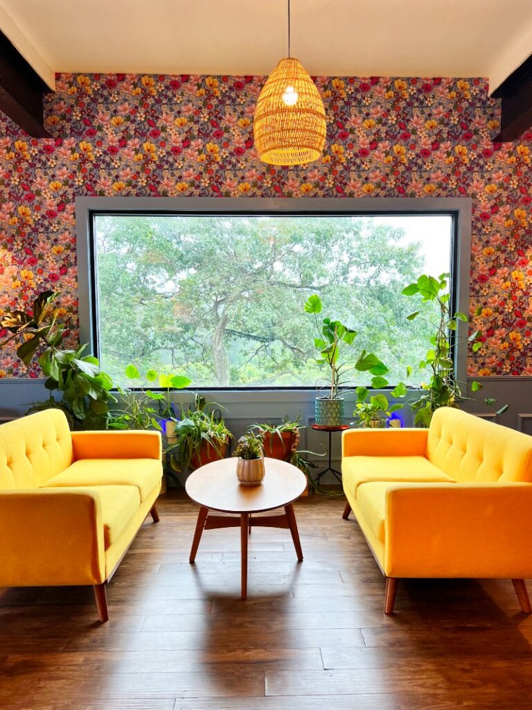 A cozy seating area in 2 Queens Coffee with yellow sofas, a round wooden table, vibrant wallpaper, and indoor plants by a large window.