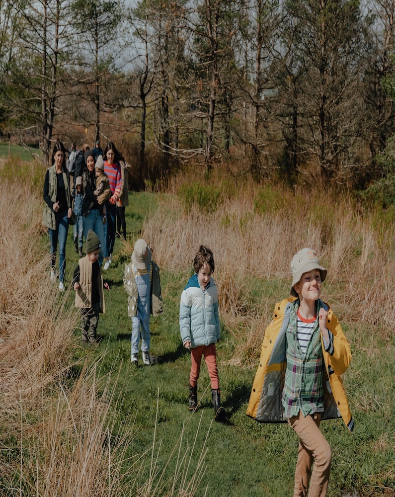 A group of children and adults walking along a grassy path surrounded by trees and tall grass on a sunny day.