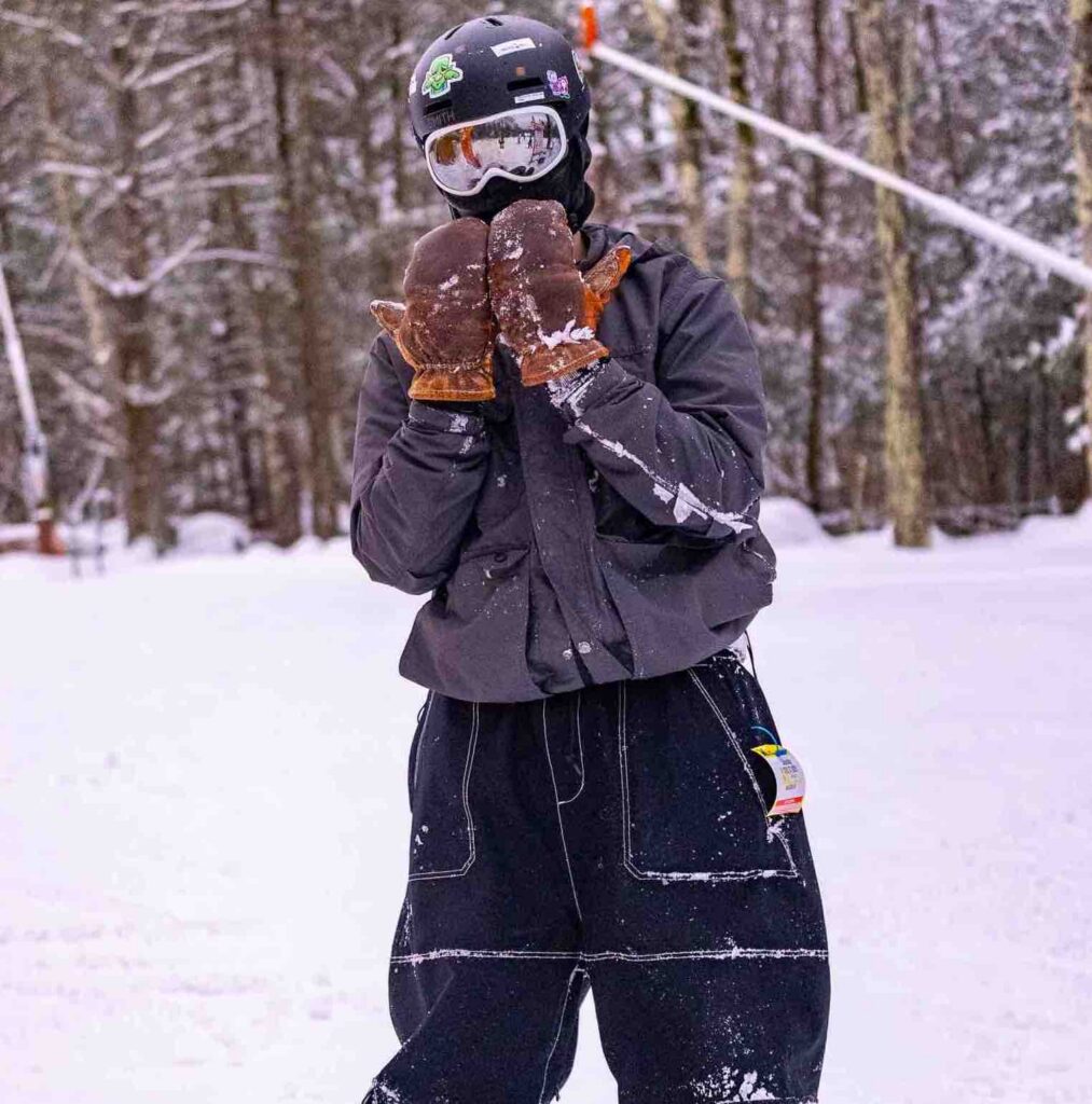 A person in winter attire stands in a snowy landscape, wearing gloves and a helmet, with snow-covered trees in the background.