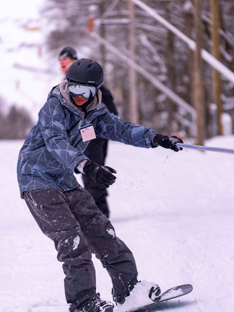 A young person snowboards down a snowy slope, dressed in winter attire, while another individual looks on in the background.