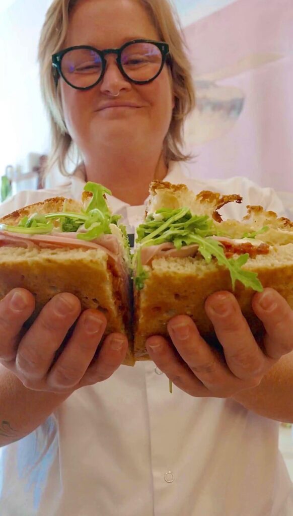 A person holds a large sandwich filled with greens and deli meats, wearing glasses and smiling while in a bright, cheerful setting.