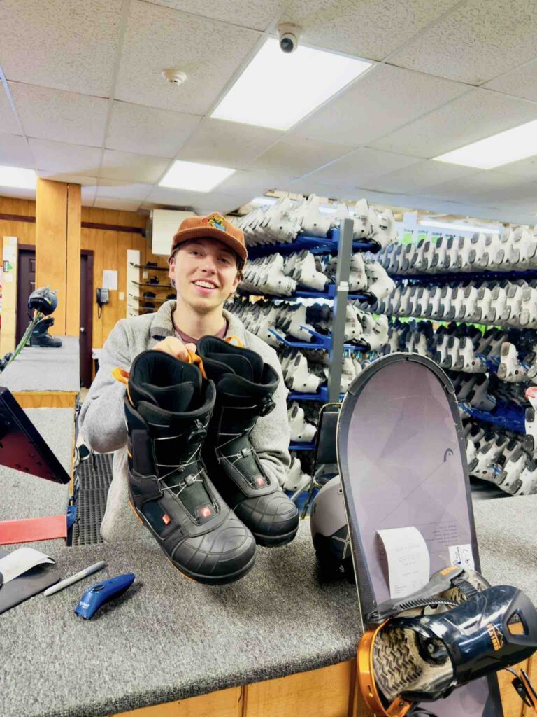 A smiling individual holding a pair of snowboarding boots in a rental shop, with snowboards visible in the background.
