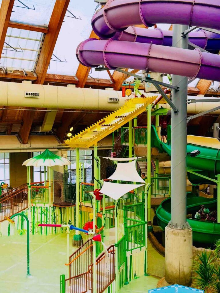 A vibrant indoor water park featuring colorful water slides, play structures, and interactive water features, surrounded by a spacious wooden ceiling.