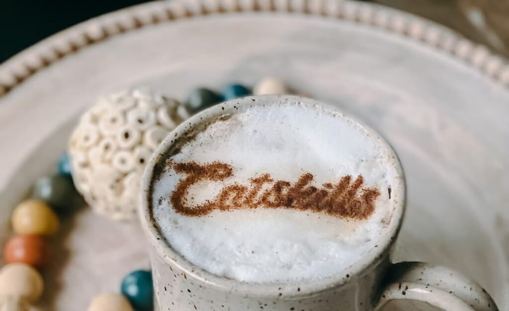 A cappuccino with the word 'Catskills' artfully sprinkled in cocoa on top, surrounded by decorative beads and a textured plate.