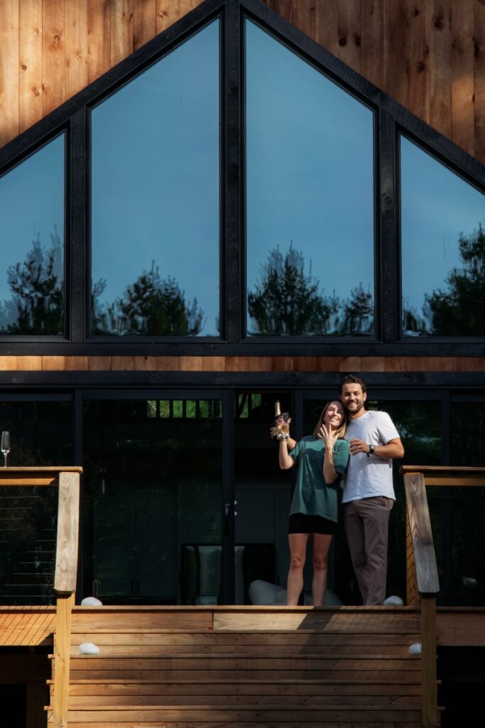 A couple stands on a wooden deck in front of a modern, glass-fronted A-frame house inteh Sullivan Catskills, smiling and holding drinks against a backdrop of trees.