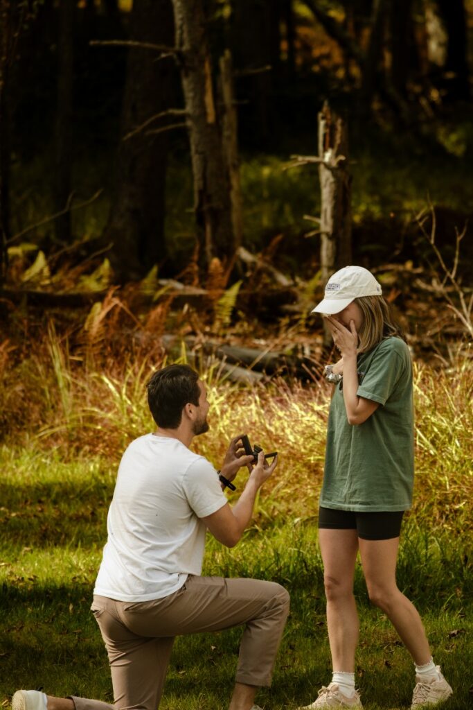 A couple in a natural setting in the Sullivan Catskills, with a man kneeling and proposing to a surprised woman wearing a cap.