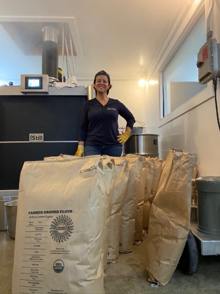 A smiling woman in gloves stands in a bright workspace surrounded by large bags, likely of coffee or other grains, with various equipment in the background.