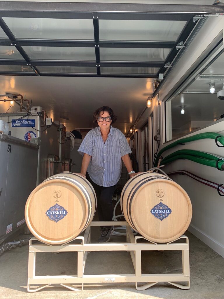 A person stands between two wooden barrels in a winery, with modern equipment and a spacious environment visible around them.