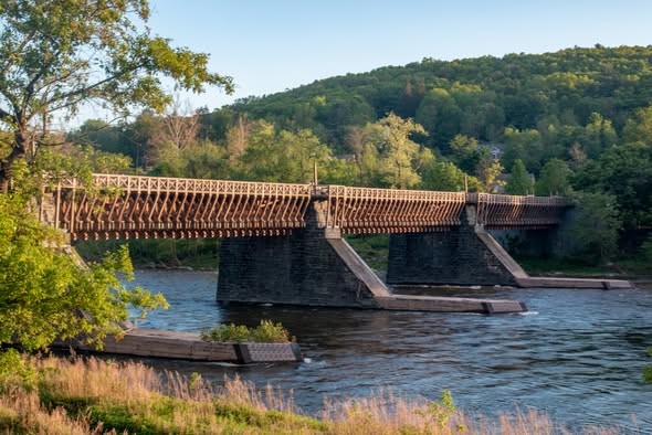 Roebling's Delaware Aqueduct arches over a calm river, surrounded by lush greenery and rolling hills, bathed in the warm glow of a setting sun.