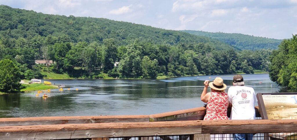 A couple enjoys a scenic view of a river surrounded by lush greenery and mountains, with a clear blue sky overhead.