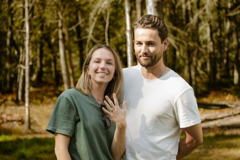 A happy couple stands together outdoors in a forest, smiling. The woman shows an engagement ring while the man poses beside her.
